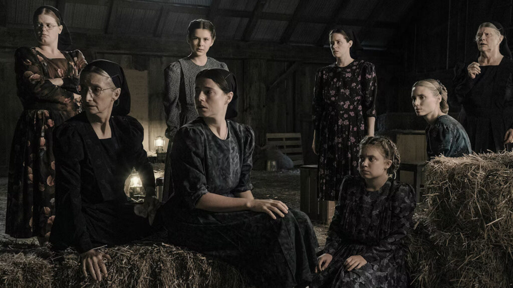 A group of women are in a barn, some are standing and some are sitting on hay bails.